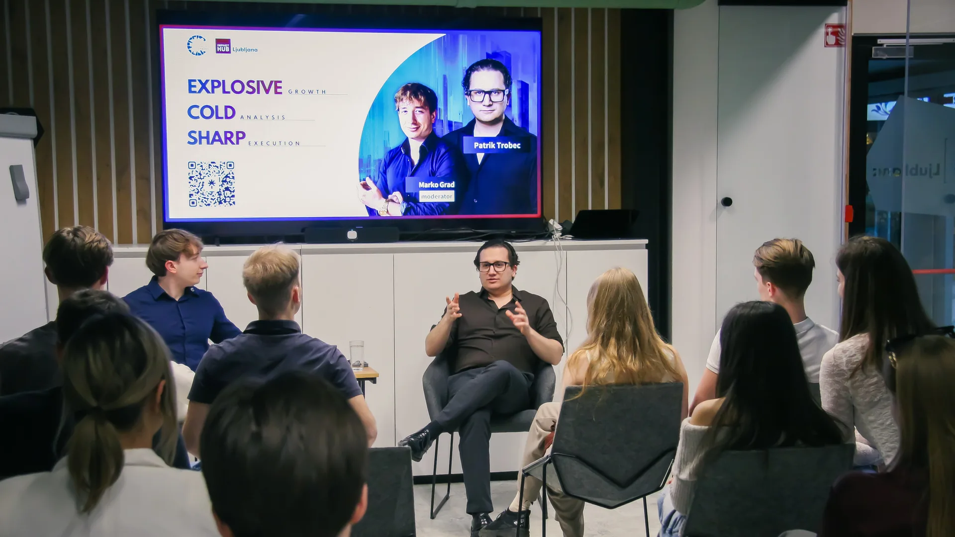 Guest Patrik Trobec speaking from a chair on the small stage at Impact Hub Ljubljana, the title slide “Explosive growth, cold analysis, sharp execution” on the screen behind him and the audience watching from the front rows