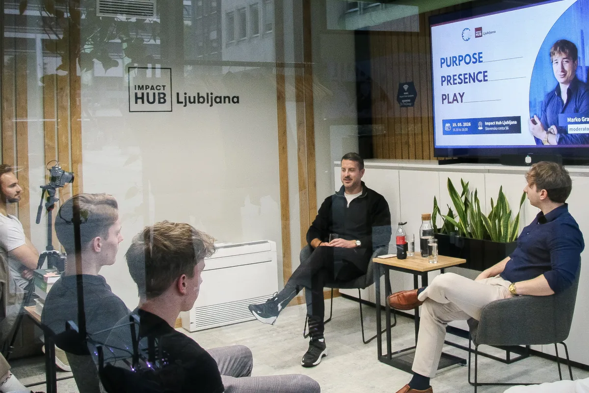 Klemen Selakovič and moderator Marko Grad seated on the stage, seen through the glass wall with the Impact Hub Ljubljana lettering, a camera on a tripod filming from the left