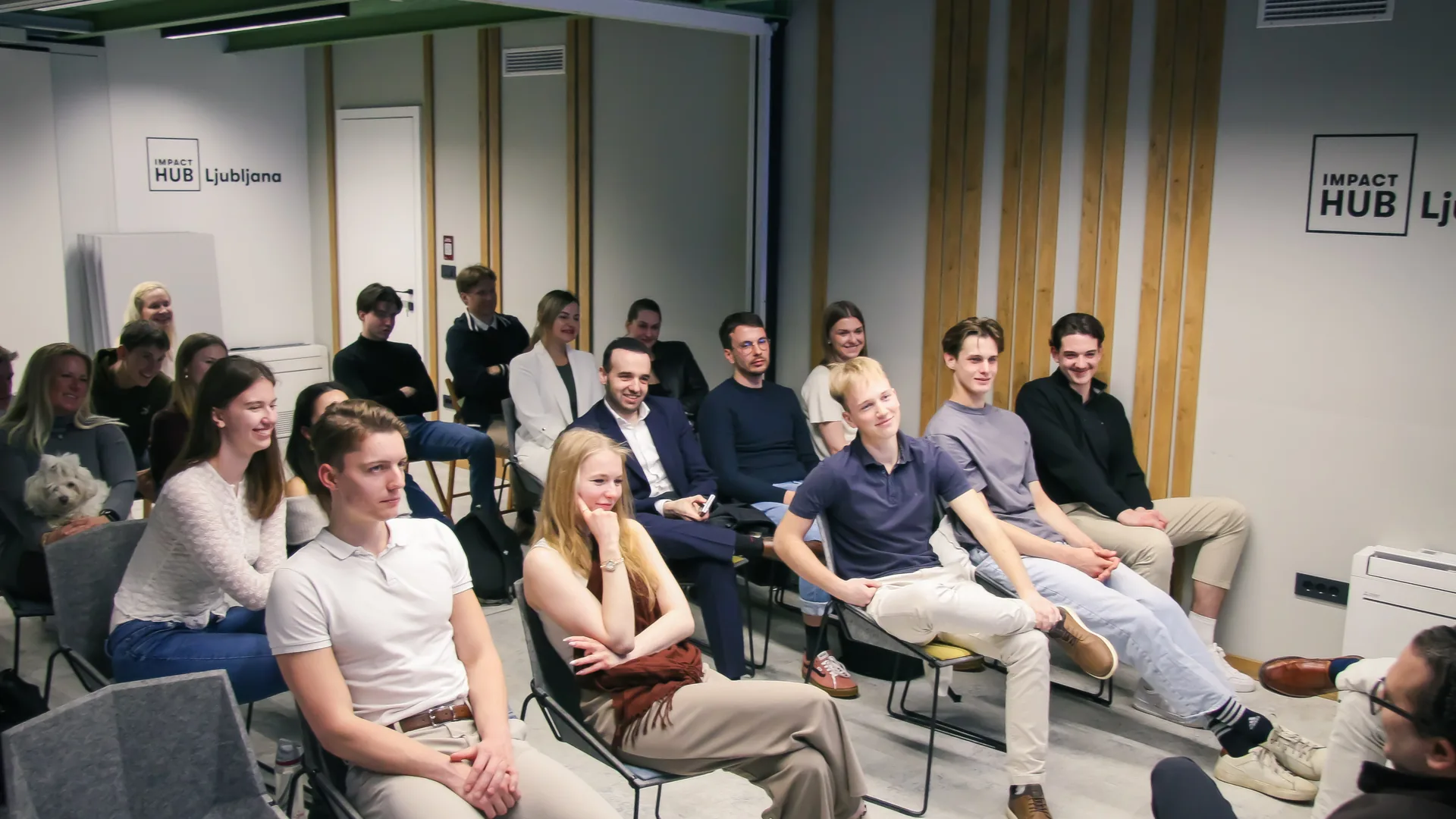 The audience at a Global Shapers talk in the Impact Hub Ljubljana event room: some twenty young people on chairs in two rows, smiling towards the stage